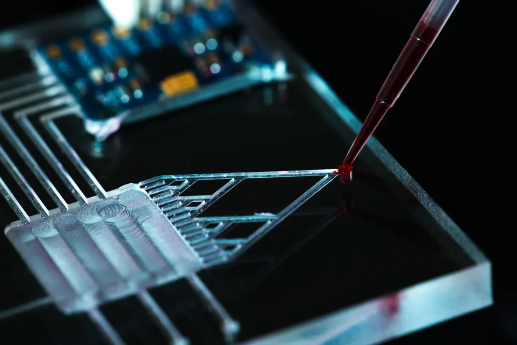 A pipette deposits a red liquid onto a transparent microfluidic chip with electronic components in the background.