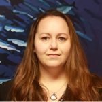 A woman with long brown hair standing in front of a blue background.