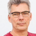 Middle-aged man with gray hair and glasses, wearing a red shirt against a white background.