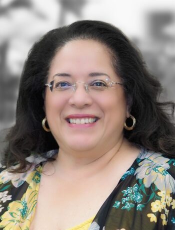 Portrait of Cyndy Luna, a smiling woman with long dark hair, wearing glasses and a floral blouse, against a blurred monochrome background. Her expression radiates experience and warmth.