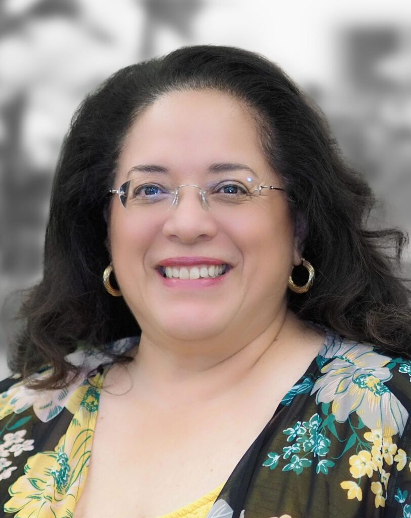 Portrait of Cyndy Luna, a smiling woman with long dark hair, wearing glasses and a floral blouse, against a blurred monochrome background. Her expression radiates experience and warmth.