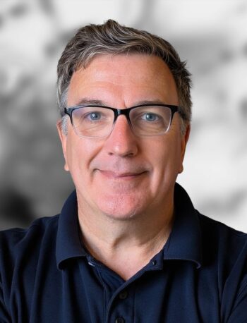 A portrait of Joe Heanue with short gray hair and glasses, wearing a navy blue collared shirt, standing in front of a blurred background.