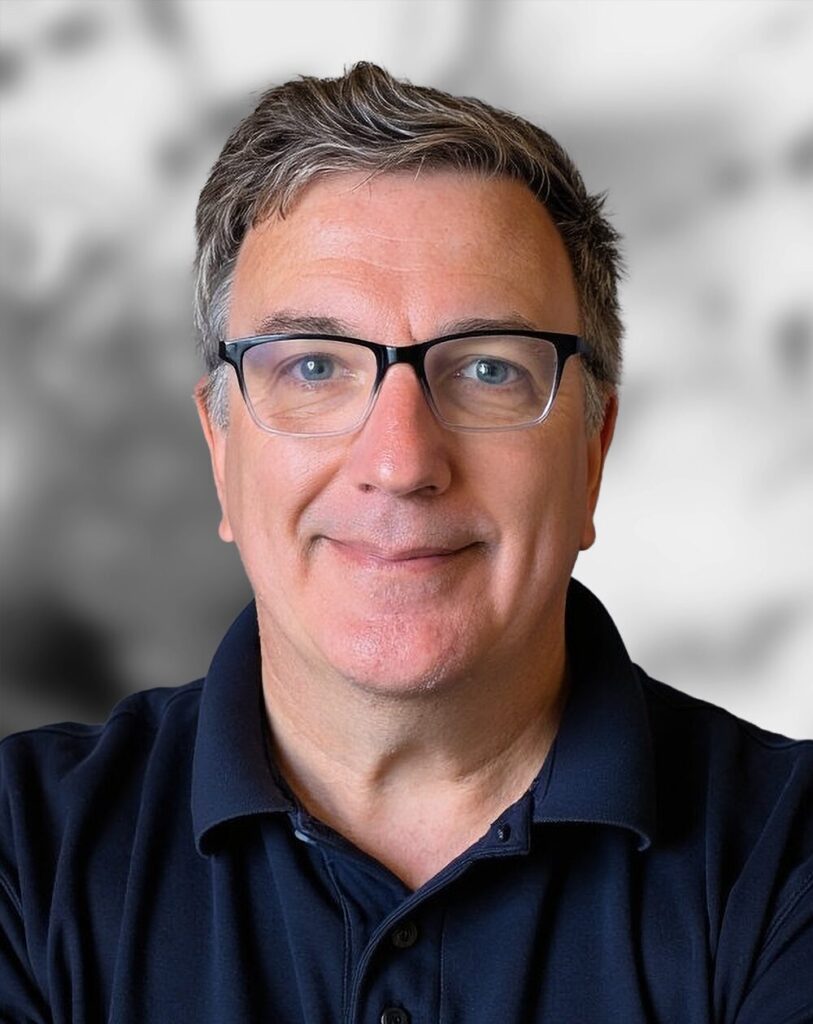 A portrait of Joe Heanue with short gray hair and glasses, wearing a navy blue collared shirt, standing in front of a blurred background.
