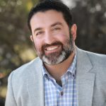 Man with a beard smiling at the camera outdoors.