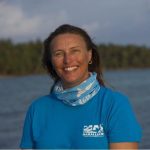 A smiling woman wearing a blue shirt standing in front of water.