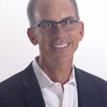 Smiling man with glasses wearing a suit and white shirt against a white background.