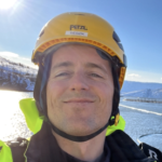 A man in a yellow helmet is smiling in front of a lake.