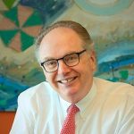 A man in glasses and a red tie smiling in front of a painting.