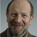 A portrait of a smiling man with a beard against a grey background.