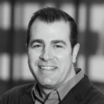 A black and white headshot of a smiling man wearing a jacket and a collared shirt.