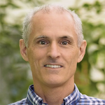 A man in a blue shirt smiles in front of plants.