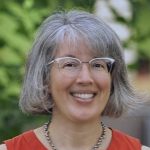 A portrait of Sheila Hemami, a smiling woman with gray hair and glasses in a red top against a blurred green background.