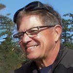 A portrait of Brad Hanson, a smiling man with glasses and a cap outdoors.