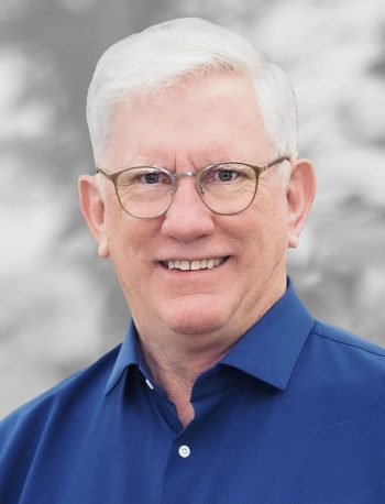 A portrait of Brian Wilfley, a smiling man with white hair and glasses in a blue shirt against a gray background.