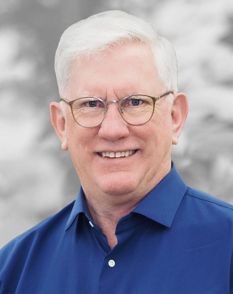 A portrait of Brian Wilfley, a smiling man with white hair and glasses in a blue shirt against a gray background.