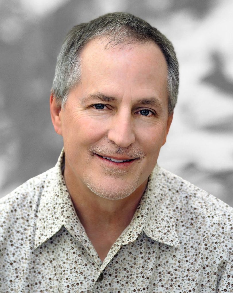 A portrait of Christopher Mitchell, a smiling man with gray hair wearing a patterned shirt.