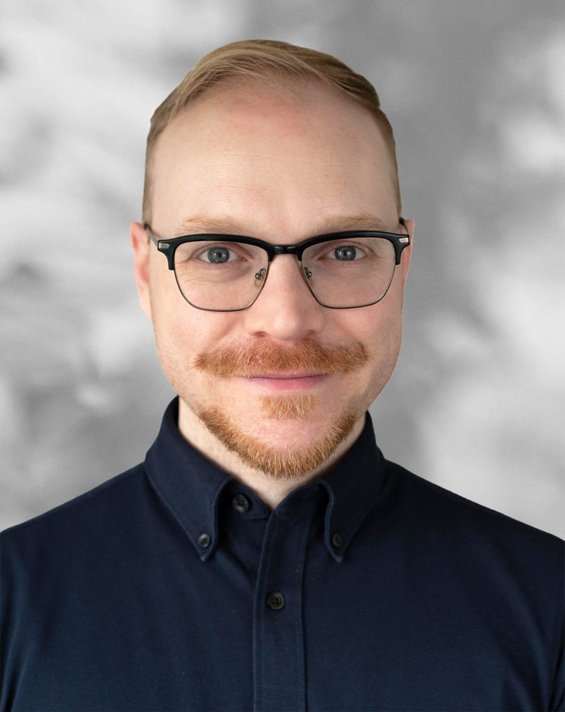 A portrait of Daniel Badali, a smiling man with glasses and a beard against a gray background.