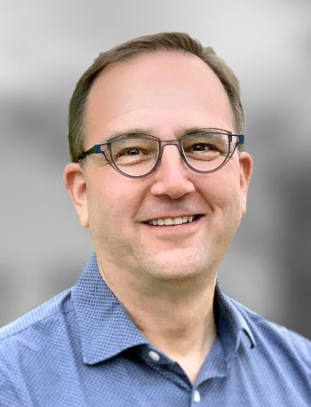 A portrait of David Shack, a smiling man wearing glasses and a blue checked shirt against a blurred background.