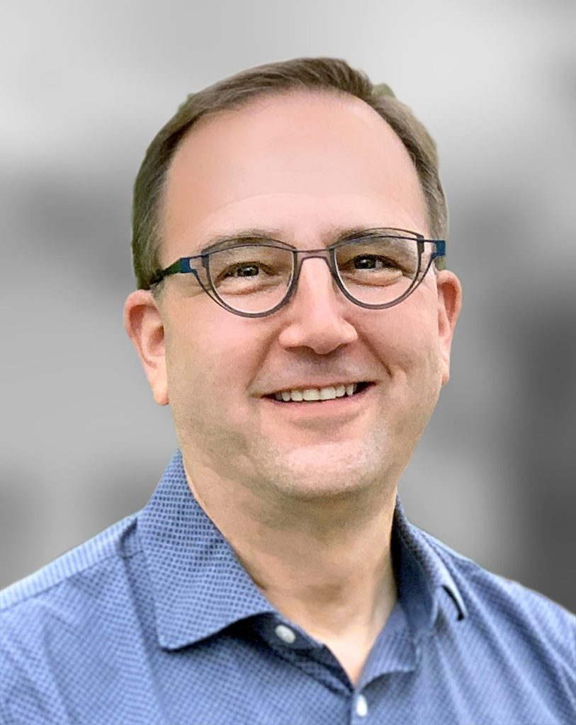 A portrait of David Shack, a smiling man wearing glasses and a blue checked shirt against a blurred background.