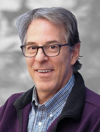 A portrait of Gene Napolitano, a smiling man with glasses wearing a checked shirt and purple fleece