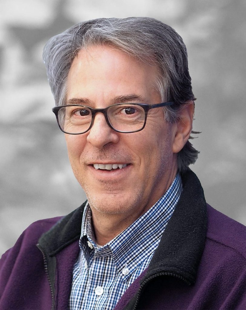 A portrait of Gene Napolitano, a smiling man with glasses wearing a checked shirt and purple fleece