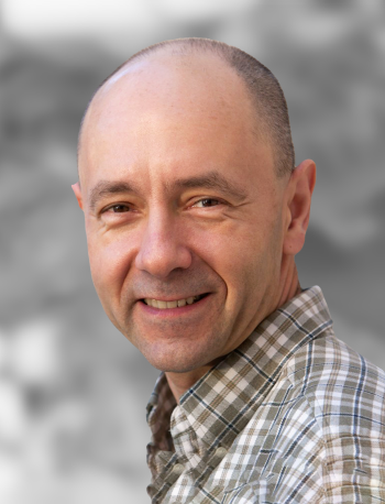 A portrait of Gregory Kovaks, a man with a smile wearing a checkered shirt against a gray background.