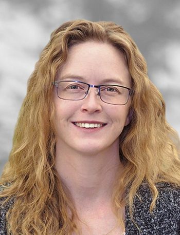 A portrait of Hailey Gryka, a woman with glasses and long curly hair smiling against a gray background.