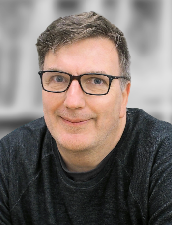 A portrait of Joe Heanue, a smiling man with glasses against a gray background.