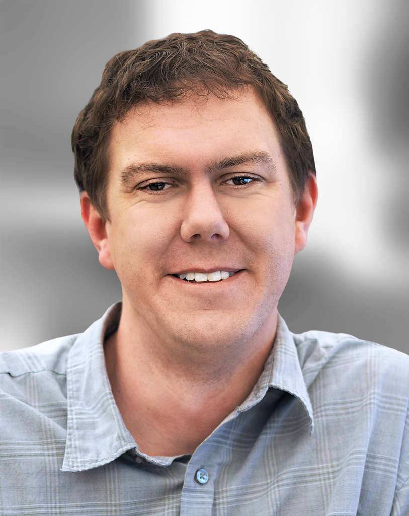 A portrait of Kevin Dunk, a smiling man with short brown hair wearing a collared, button-up shirt.