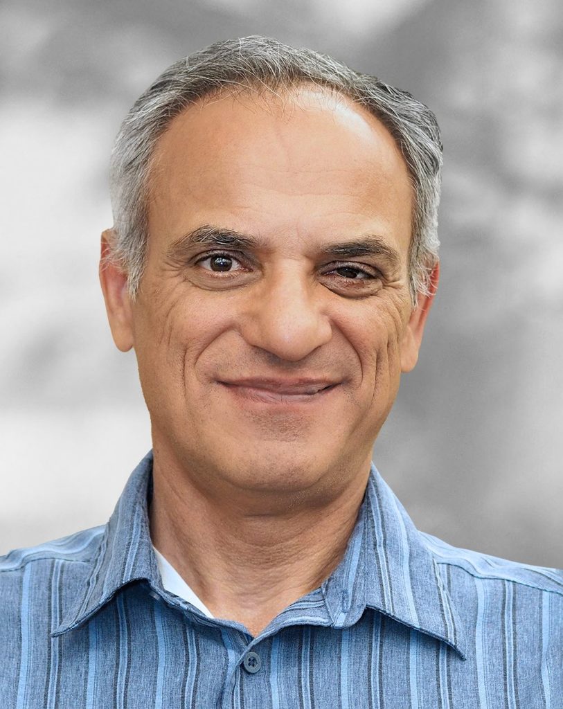 A portrait of Mansour Saleki, a smiling man with graying hair wearing a blue striped shirt against a blurred gray background.