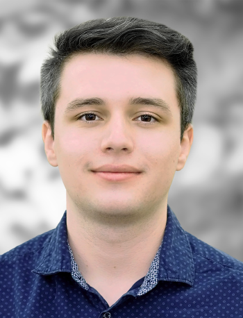 A portrait of Nicholas Mann, a man with a neutral expression, wearing a blue patterned shirt against a gray background.