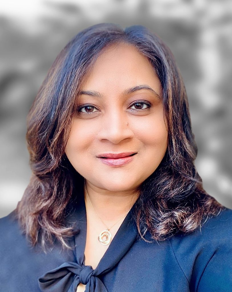 A portrait of Pooja Kartik, a woman with shoulder-length hair, wearing a black top with a bow at the collar.