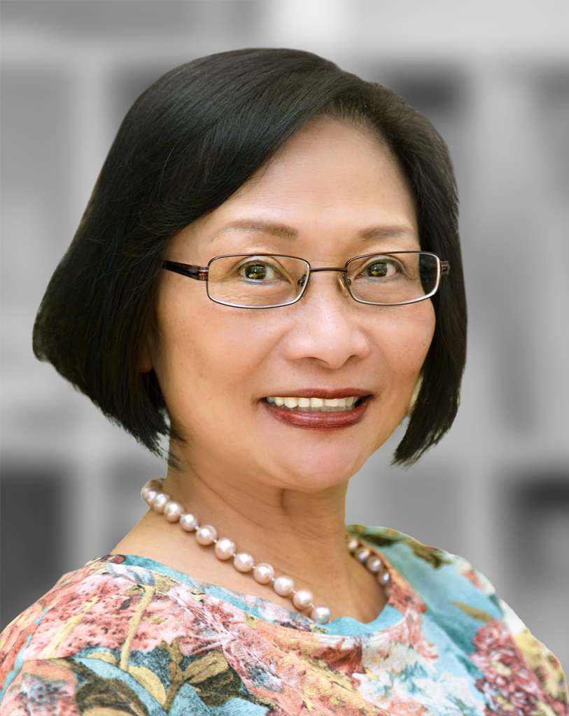 A portrait of Quan Tran, a smiling woman with short hair wearing glasses, a floral blouse, and a pearl necklace.