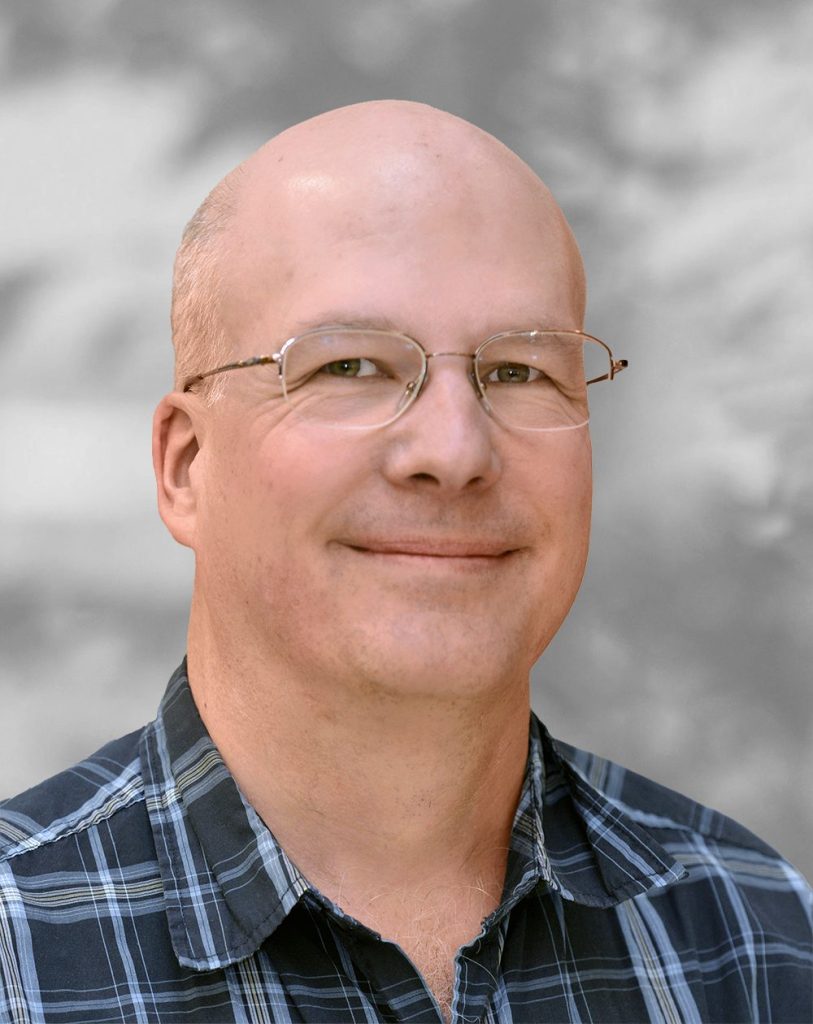 A portrait of Roger Stenerson, a smiling man wearing glasses and a plaid shirt against a blurred background.