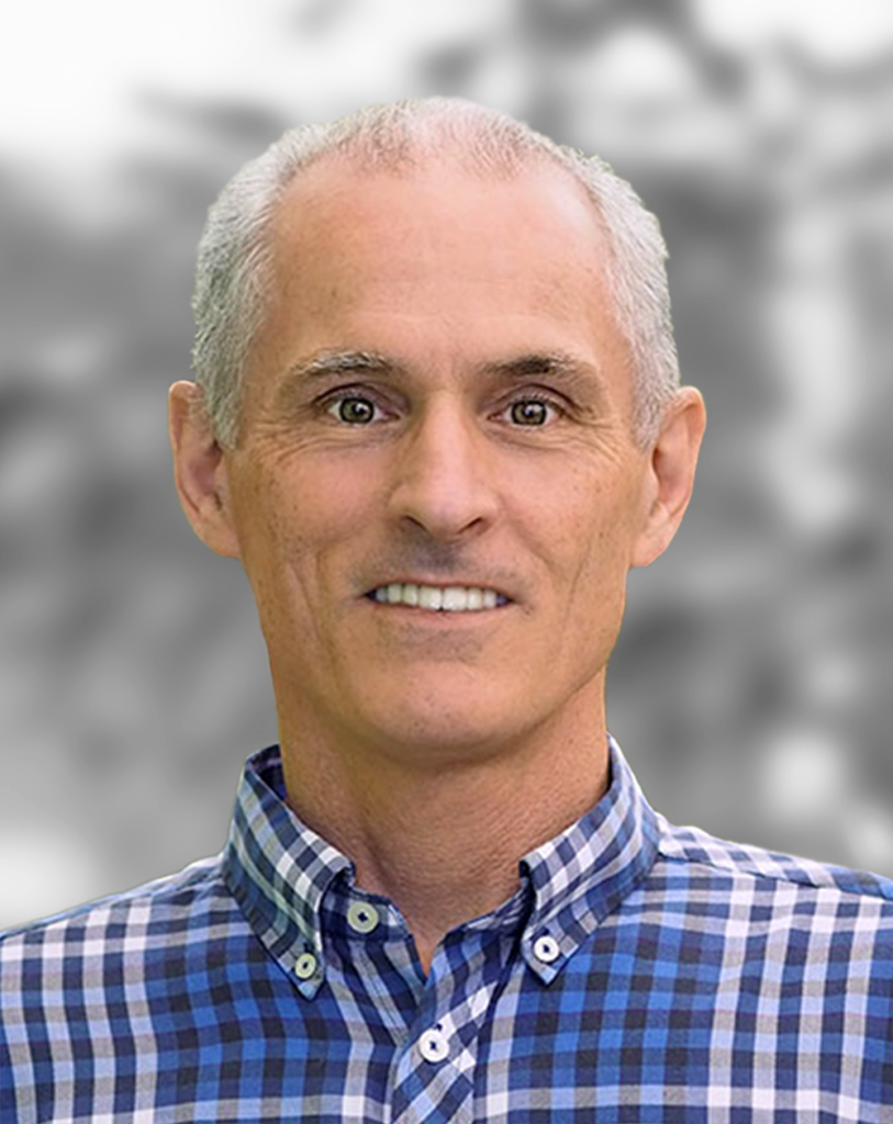 A portrait of Ryan McGuinness, a smiling man wearing a checkered shirt, against a blurred background.