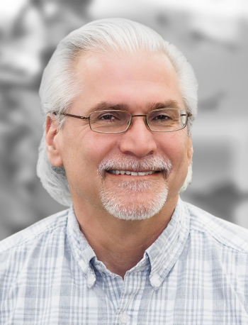 Portrait of Tachi Callas, a smiling man with gray hair and a beard, wearing glasses and a plaid shirt, set against a blurred background.