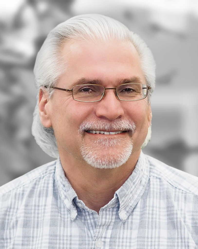 Portrait of Tachi Callas, a smiling man with gray hair and a beard, wearing glasses and a plaid shirt, set against a blurred background.
