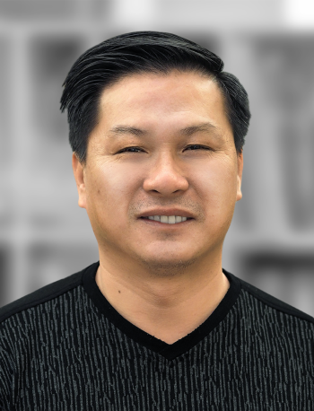 A portrait of Ty Ta, a man wearing a black shirt smiling against a blurred gray background.