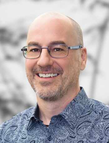 A portrait of Nick Hawson, smiling man with glasses wearing a patterned blue shirt against a blurred background.