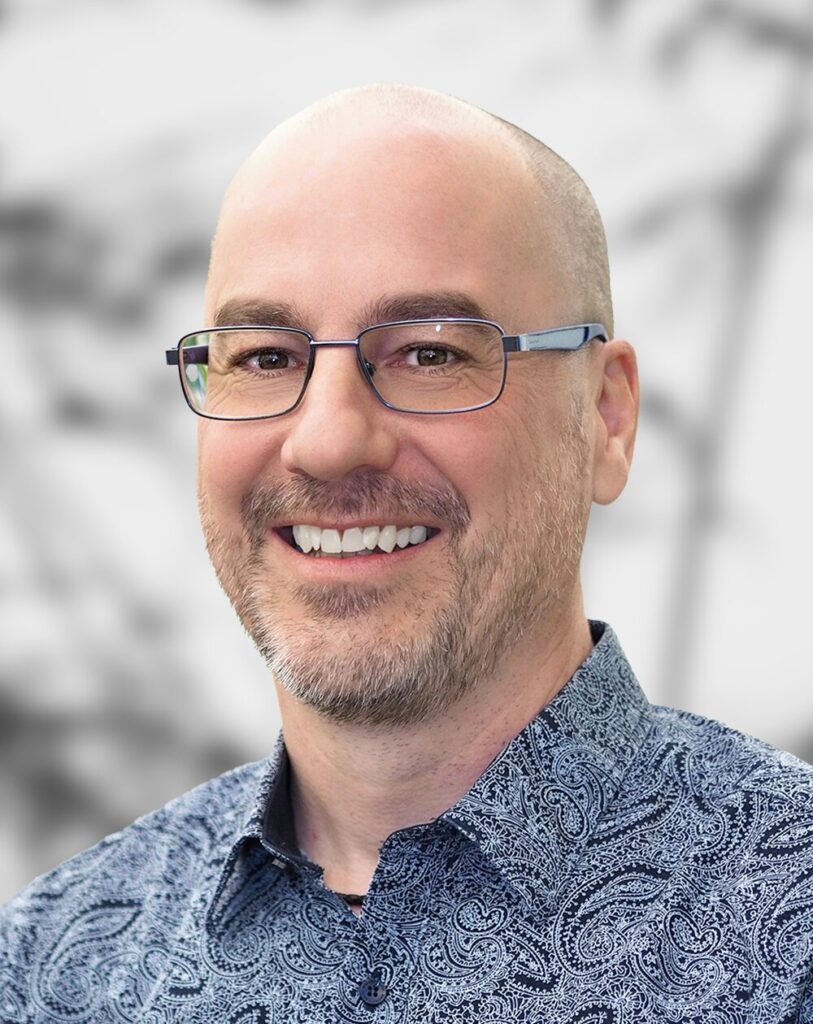 A portrait of Nick Hawson, smiling man with glasses wearing a patterned blue shirt against a blurred background.