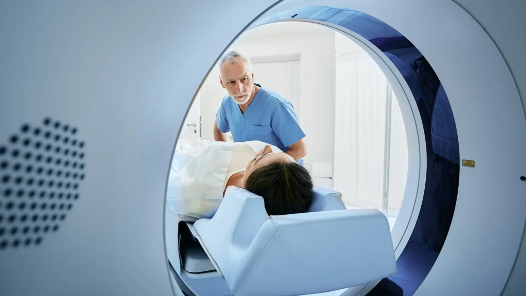 A healthcare professional assists a patient lying on a bed entering a large medical imaging machine, likely for an MRI or CT scan, in a clinical setting.