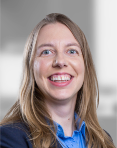 Smiling woman with long blonde hair wearing a blue shirt and dark blazer, photographed against a blurred gray background.