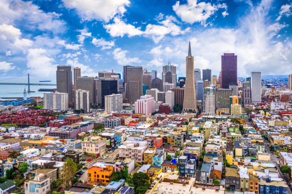Aerial view of downtown San Francisco with skyscrapers, colorful low-rise buildings, and the Bay Bridge visible in the background under a partly cloudy sky.
