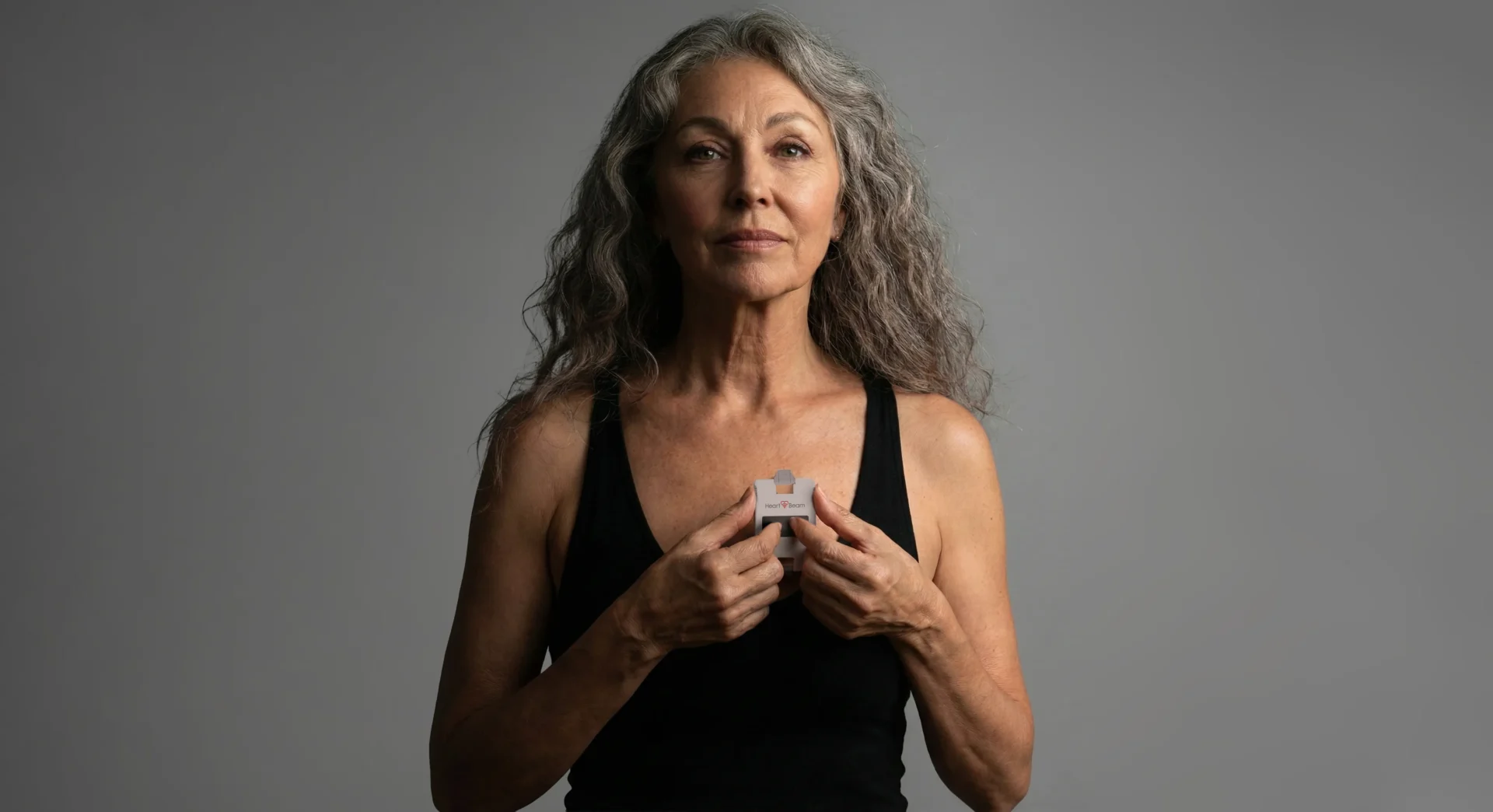 An older woman with long gray hair holds a small medical device to her chest, standing against a plain gray background.