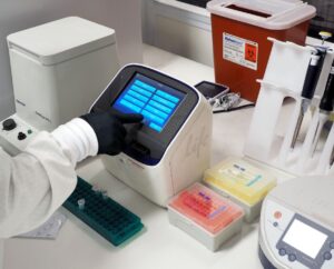 A gloved hand operates a laboratory touchscreen device surrounded by pipettes, sample containers, and lab equipment on a workbench.