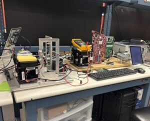 A workbench with electronic testing equipment, circuit boards, a multimeter, a keyboard, and a small monitor arranged on a blue table.