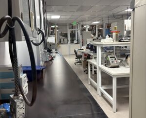 A laboratory workspace with scientific equipment, white tables, chairs, and storage boxes along a clean, organized corridor.