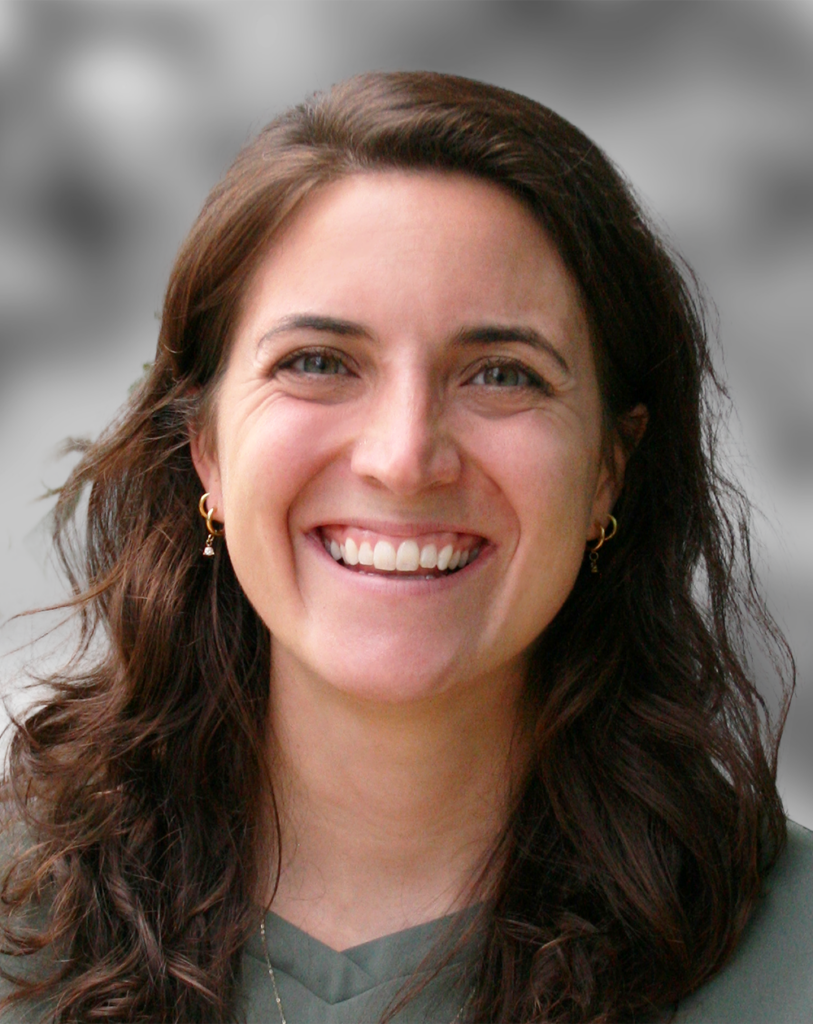 A photo of Anne Preut, a woman with wavy brown hair smiling, wearing a green top and small gold hoop earrings, against a blurred gray background.
