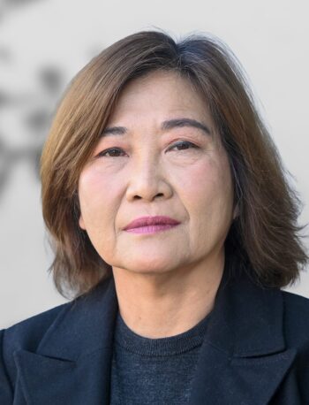 A photo of Gertrudes Armena, a woman with shoulder-length brown hair, wearing a black jacket and dark sweater, looking directly at the camera with a neutral expression against a plain background.