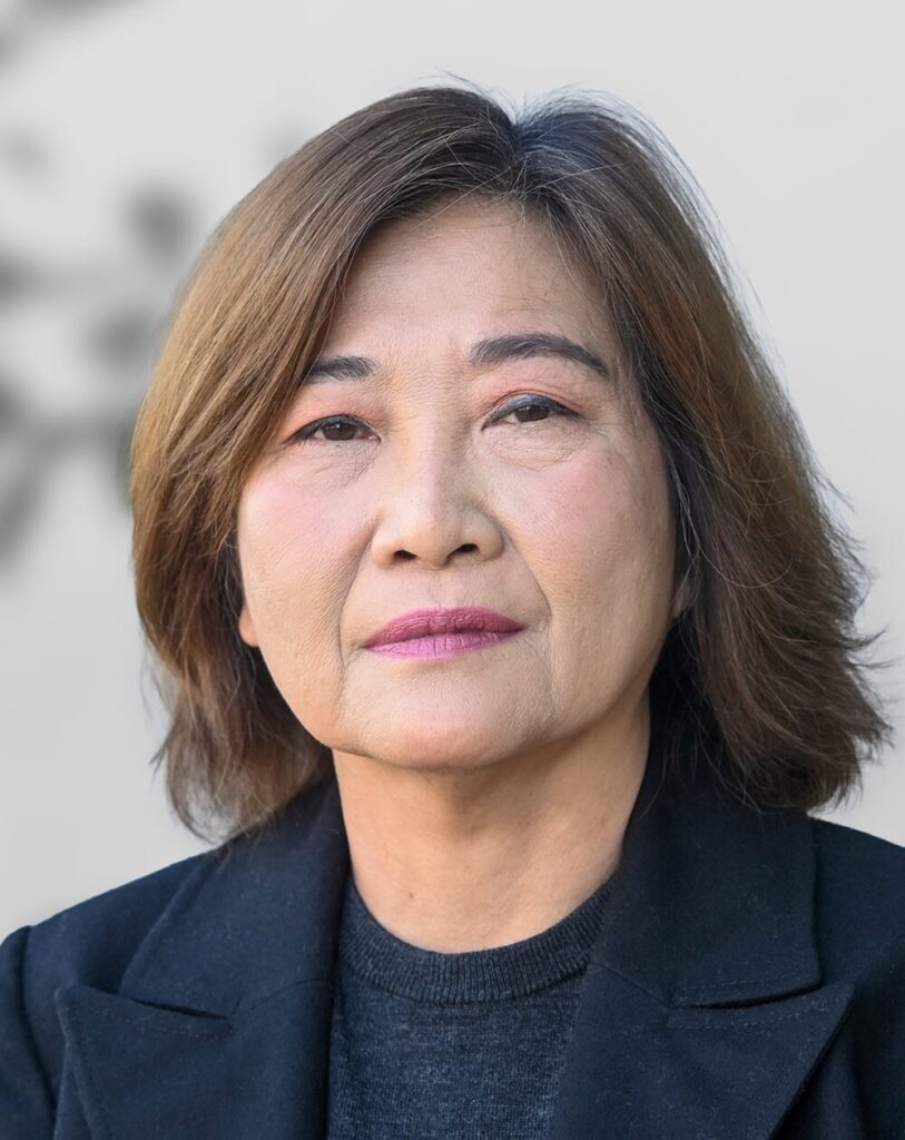A photo of Gertrudes Armena, a woman with shoulder-length brown hair, wearing a black jacket and dark sweater, looking directly at the camera with a neutral expression against a plain background.